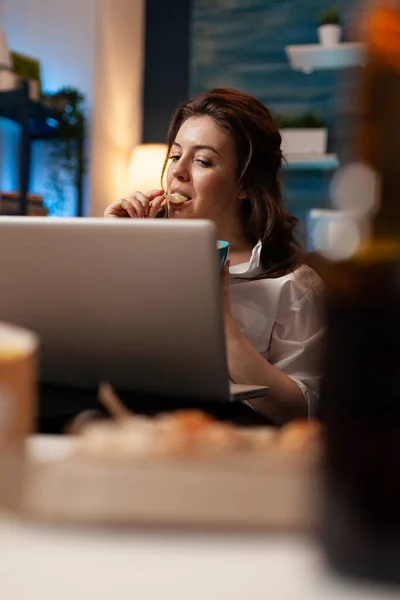 Mujer relajante en el sofá comiendo sabroso bocadillo mientras ve la película de comedia en el ordenador portátil — Foto de Stock