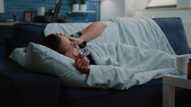 Крупный план больного человека с оксиметром на пальце, лежащем на диване — стоковое видео