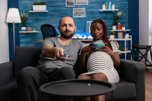 Pregnant interracial couple looking at camera watching television — Stock Photo, Image