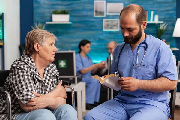 Caregiver support nurse reading book stories to senior disabled pensioner — Stock Photo, Image