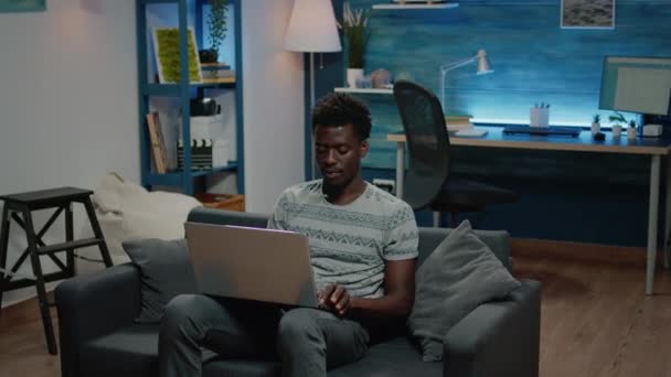 Afrykański amerykański freelancer za pomocą laptopa do zdalnej pracy podczas siedzenia na kanapie — Wideo stockowe
