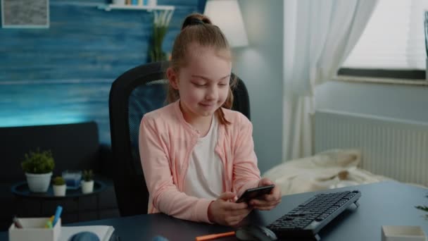 Маленькая девочка смотрит на дисплей смартфона и улыбается за столом — стоковое видео