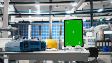 Yeşil ekranlı bir güneş enerjisi üretim tesisinde boş mühendislik masası. Robot kollar ve güneş paneli montaj hatlarıyla çevrili endüstriyel çalışma alanı. Performans izleniyor.
