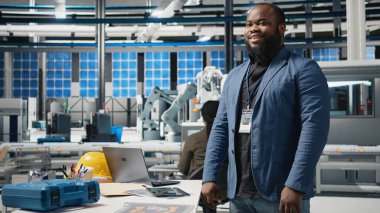 Üniforma içindeki siyah usta mühendis, yeşil enerjinin geleceğinde teknik hassasiyet ve kurumsal stratejiyi temsil eden güneş paneli üretimini onaylıyor. Montaj hattı. Kamera A.