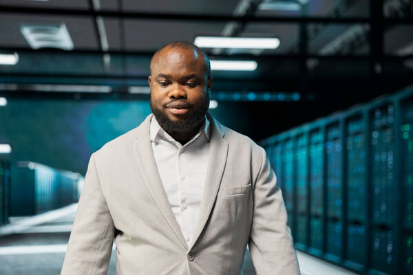 Admin walking in high tech data center, doing checkup to prolong hardware life span and avoid downtime. African american server hub IT specialist monitoring server infrastructure rigs