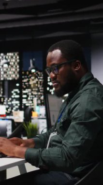 Modern çalışma alanındaki dikey Video Siyah Erkek Yöneticisi bir bilgisayar monitöründe öngörü ve iş stratejisini analiz ederek, gece geç saatlerde üretkenlik için rapor yazmayı ve plan yapmayı güvence altına alır