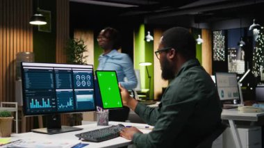 Genç siyahi işadamının verileri incelediği tabletin yanındaki yeşil ekranda, analitik raporlar üzerinde çalışarak küresel genişleme ve iş yönetimi görevleri için çözümler bulmaya çalışıyor. Kamera A.