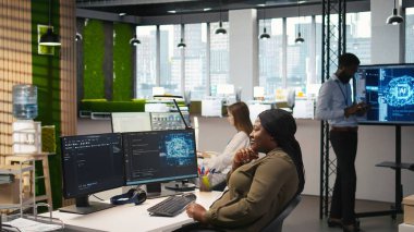 Başlangıç ofisindeki bilgisayar bilimci yapay zeka sistem ölçümlerini gözden geçiriyor, performans gösterge panellerini izliyor. Afrika kökenli Amerikalı bir kadın, bilgisayar ve kamera A 'daki araçları kullanarak sinirsel ağları ayarlıyor.