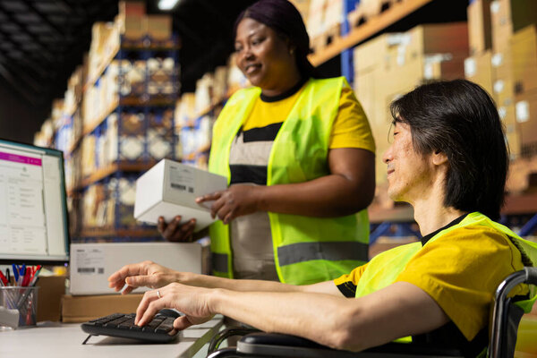 Industrial worker with disability uses airway bill numbers for tracking info on software, monitoring the order parcel status in a logistics hub with a colleague. Managing shipment operations.