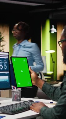 Tabletin üzerindeki dikey video yeşil ekran, verileri inceleyen, analitik rapor yazma ve küresel genişleme ve iş yönetimi görevleri için çözümler bulma üzerinde çalışan genç siyahi iş adamlarının yanında