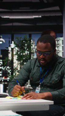 Dikey Video Siyah iş adamı, yeni bir geliştirme görevini hatırlamak için yapışkan kağıt etiketi kullanarak monitöre yapışkan bir not bırakıyor. Geceleri karanlık ofis, verimlilik motivasyonu. Kamera A