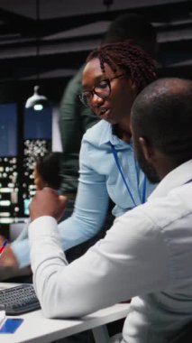 Dikey Video İki siyahi işçileri masadaki sanal bir asistanla etkileşime geçtiler, yapay zeka ve akıllı otomasyon ile çevrimiçi iletişimi geliştirdiler. Veri güdümlü yapay zeka çözümleri. Kamera.