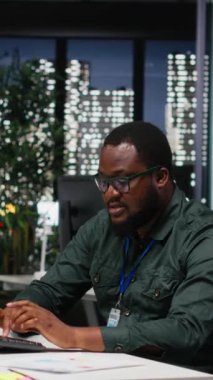 Dikey Video Afrikalı Amerikalı Danışmanı, geceleri karanlık bir ofiste karmaşık bir soruna odaklanarak, masaüstü seçenekleri tasarlıyor ve öngörüyor. Dikkatli sonuç çıkararak mantıklı bir çözüm arıyorum.