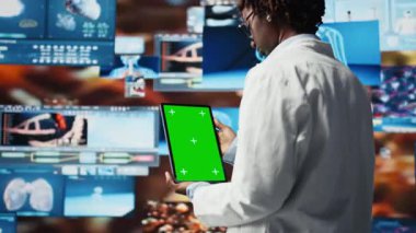 Klinisyen tarafından kullanılan yeşil ekran tableti multimedya duvarındaki tıbbi raporlara bakıyor. Laboratuvar önlüğü giyen doktor model cihazının tıbbi araştırma verilerini analiz ediyor. Kontrol panelleri, kamera A.