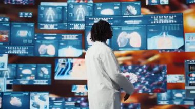Kolları çapraz olan klinisyen multimedya duvarındaki tıbbi raporlara bakıyor. Önlük giyen doktor tıbbi araştırma verilerini analiz eder, etkileşimli gösterge panellerini izler, kamera A