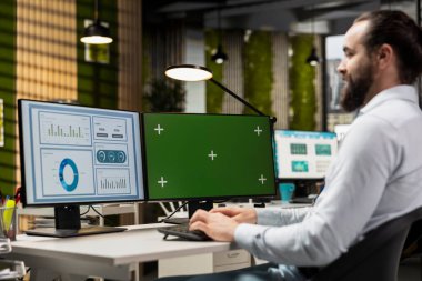 Genç iş analisti monitörde kroma anahtarıyla hava tahminlerini inceliyor, brifing için performans ölçümleri üzerinde çalışıyor. Küresel genişleme ve ağ oluşturma için rakamlar artırılıyor.