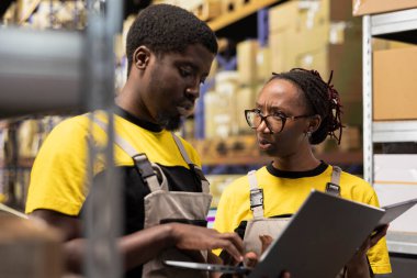 Depo çalışanları paketleri denetliyor dizüstü bilgisayardaki bilgileri takip ediyor, giden sevkiyatlar için paletler hazırlıyor. Depo rafları, karton kutular ve envanter işleme araçlarıyla organize depo.