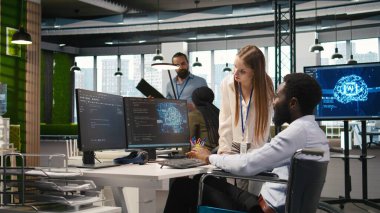 Küçük Dev ve tekerlekli sandalyedeki iş arkadaşı yazılım uygulamaları geliştiriyor, kodu optimize ediyor. Belden aşağısı felçli kadın teknisyen ve meslektaş, A kamerasındaki darboğazları çözmek için yapay zeka kullanıyor.