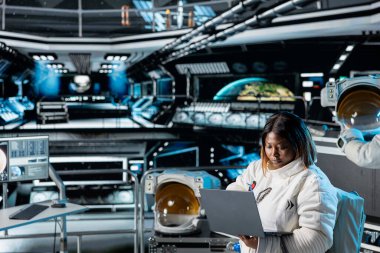 Dişi astronot bilim adamı uzay aracı kontrol arayüzünü dizüstü bilgisayarla kullanıyor. Uzay gemisinde kozmonot kıyafeti giyen kadın araştırmacı araştırma yapıyor, sistem teşhislerini izliyor.