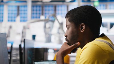 Siyah mühendis otomatikleştirilmiş fabrikada güneş yazılımı ayarları üzerinde çalışarak üretim sürecini optimize ediyor. Dijital araçlar ve endüstriyel robotlar kullanılarak optimal sistem yapılandırmasını sağlar. Kamera A.