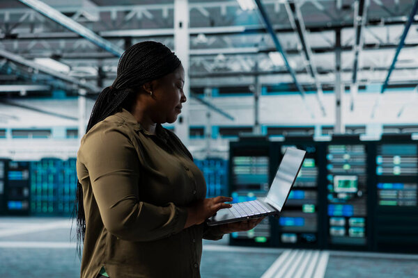 Woman in data center conducting tests on infrastructure using software to fix bugs. African american server hub worker using laptop to refine algorithms for faster processing, updating core services
