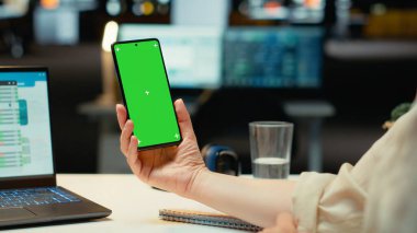 Ofis çalışanı masasında krom ekranlı akıllı telefonunu tutuyor, mesai saatlerinden sonra kurumsal çalışma alanında görevleri çözüyor. Kadın çalışan girişimci yönetiminde çalışıyor. Kamera B.