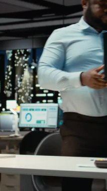 Dikey Video Afrikalı Amerikalı iş adamı ofiste duruyor ve çözümler arıyor, düşünceli oluyor ve şirket büyümesi için yeni fikirler buluyor. Gece geç saatlerde görevleri öngören bir danışman. Kamera.