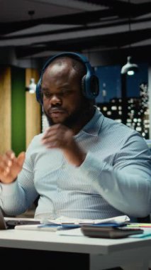 Dikey Video Siyah iş adamı müzik dinler ve üst vücut kaslarını esneterek karmaşık işleri bitirmek için gece geç saatlere kadar çalışır. Kulaklıkları bırakıp işini bitiriyor. Kamera.