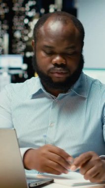 Dikey Video Afrikalı Amerikalı adam el yazısı özeti hazırlıyor ve ofiste saatler sonra güncellemeler yapıyor. Günlük planlama, iş hedefleri ve gündem belgelerinin sağlanması. Kamera B.