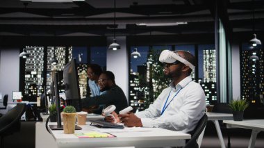 Genç siyah işadamı sanal gerçeklik kulaklığı ve diğer cihazları kullanarak bir strateji oluşturuyor, öngörülerde bulunuyor ve başarı için risk değerlendirmesini belgeliyor. İş hedefleri.