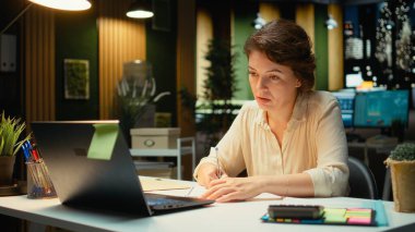 Şirket çalışanı masa lambasının altına detaylı bir rapor yazmaya odaklanmış. Güncellemeleri özetlemek, gündemi düzenlemek ve şirket yönetim planları için içgörü taslakları hazırlamak. Kamera A.