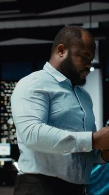 Dikey Video Afrikalı Amerikalı iş adamı ofiste duruyor ve çözümler arıyor, düşünceli oluyor ve şirket büyümesi için yeni fikirler buluyor. Gece geç saatlerde görevleri öngören bir danışman. Kamera.