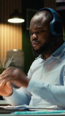 Dikey Video Siyah iş adamı müzik dinler ve üst vücut kaslarını esneterek karmaşık işleri bitirmek için gece geç saatlere kadar çalışır. Kulaklıkları bırakıp işini bitiriyor. Kamera.