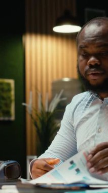 Dikey Video Siyah erkek çalışanı, çalışma saatleri dışında lambasını ve laptopunu kapatıyor, teslim tarihlerine yetişmek ve hedefe ulaşmak için gece geç saatlere kadar çalışıyor. Fazla mesai yapıyor ve strateji geliştiriyor.