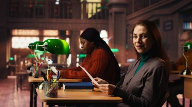 Olgun bir kadın, yeşil lamba altında bir halk kütüphanesinde verileri inceliyor. Belgeleri okumaya ve okumaya son derece odaklanmış durumda..