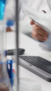 Dikey video laboratuarı profesyoneli, şeffaflık ve tutarlılık için kontrol altında test tüpü örneğini inceler. Bilim adamı sıvı özelliklerini gözlemlemek ve tespit etmek için lamba aydınlatmasını kullanır.