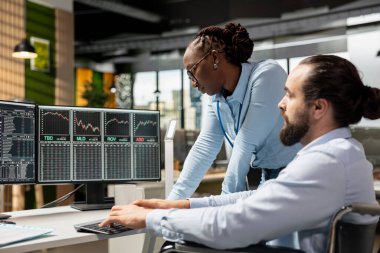 Risk analisti ve kapsayıcı ofiste engelli iş arkadaşı borsa değişkenliğini incelemek için ticaret platformunu kullanıyor. Tekerlekli sandalyedeki engelli kişi ve iş arkadaşı portföy raporlarını analiz ediyor.