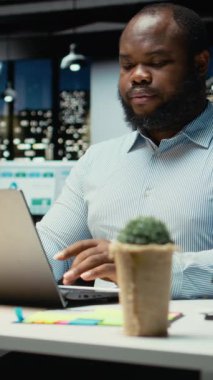 Siyah iş adamının dikey video portresi, gece ofiste oturup not defterine veri yazmadan önce eleştirel düşünme ve düzeltme yapıyor. Önemli fikirlerin fazla mesai olarak belgelenmesi