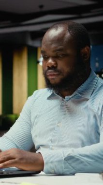 Dikey Video Afrikalı Amerikalı yorgun adam ofiste saatler sonra uyanık kalmaya çalışıyor, çok çalışıyor ve işi bitirmek için çabalıyor. Saatler sonra kendini çok yorgun ve bunalmış hisseden kişi, uykusuzluk