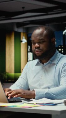 Dikey Video Afrikalı Amerikalı adam gece ofiste oturup not defterine veri yazmadan önce eleştirel düşünme ve düzeltme yapıyor. İşçi önemli fikirleri belgeliyor. Kamera.