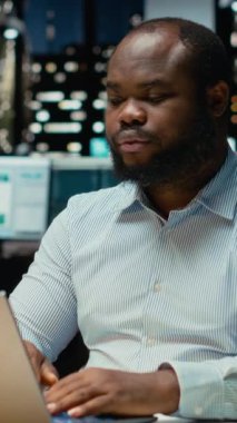 Dikey Afrikalı Amerikalı erkek işçi sırt ve boyun kaslarını esnetiyor, kas gevşemesi ve gevşemesi için duruşunu düzeltiyor. Ofisteki yoğun hareketlilikten dolayı gergin ve boyun tutulması