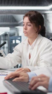 İş yerinde Asyalı iş arkadaşının yanındaki gösterge çözeltisiyle dolu laboratuvar camı kullanan dikey video kimyacısı. Laboratuvar bilimcisi bilgisayarını kullanarak kimyasal sıvıyla silindiri inceliyor.