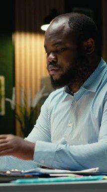 Dikey Video Afrikalı Amerikalı adam piyasa verilerini gözden geçiriyor ve iş hedefleri için strateji geliştiriyor, dizüstü bilgisayardaki hesap tablolarını düzeltme için kullanıyor. Çok uluslu şirkette saatler sonra fazla mesai. Kamera.