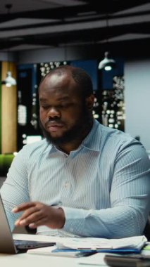 Dikey Video Afrikalı Amerikalı adam piyasa verilerini gözden geçiriyor ve iş hedefleri için strateji geliştiriyor, dizüstü bilgisayardaki hesap tablolarını düzeltme için kullanıyor. Çok uluslu şirkette saatler sonra fazla mesai. Kamera.