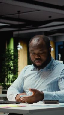 Dikey Video Siyah iş adamı cep telefonundan iç çekip mesajları kontrol ediyor, modern ofisteki bir tebligat yüzünden dikkati dağıldıktan sonra tükenmiş ve kaşlarını çatmış hissediyor. Geceleri işyeri sorunları