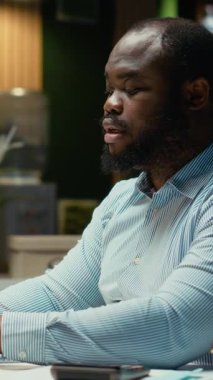 Dikey Video Afrikalı Amerikalı adam diğer masalara bakıyor ve ofiste yalnız olduğunu fark ediyor, fazla mesai yapıyor ve tükenmişlerle uğraşıyor. Uykucu aşırı yüklenmiş kişi gece geç saatlere kadar çalışıyor.