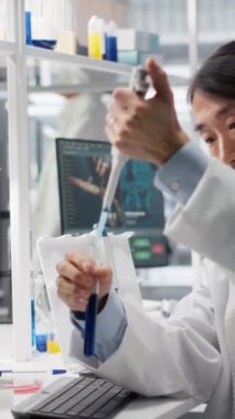 Dikey video laboratuvar mühendisi kimyasal reaktörle pipetleme işlemi yapıyor, reaksiyon testleri için çözümleri karıştırıyor. Kıdemli bilim adamı sıvıyı test tüpüne aktarmak için pipetini kullanıyor, karıştırmak için sallanıyor.