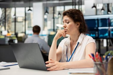 Beyaz kadın yönetici cihaz üzerinde analiz yapıyor, şirket bilgileriyle ilgili karışık bir ifadeyle finansal verileri analiz ediyor. Endişeli kadın, çenesi üzerinde laptopta belgeleri gözden geçiriyor.