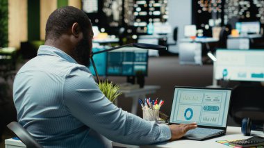 Erkek ofis çalışanı geceleri rakamları ve mali veri ölçümlerini kontrol ediyor, güncellemeleri belgeliyor ve yaklaşan iş etkinliklerinin gündemini düzenliyor. İdari görevler.