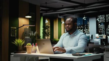 Afrika kökenli Amerikalı iş adamı işlerini bitiriyor ve masa lambasını kapatıyor. Kompleks proje için fazla mesai yaptıktan sonra gece geç saatlerde ofisten çıkıyor. İşi bitirdikten sonraki zamanı kontrol ediyorum. Kamera A.